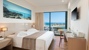 A hotel room with a bed, a desk, a chair, a TV, and a large window with a view of the sea.
