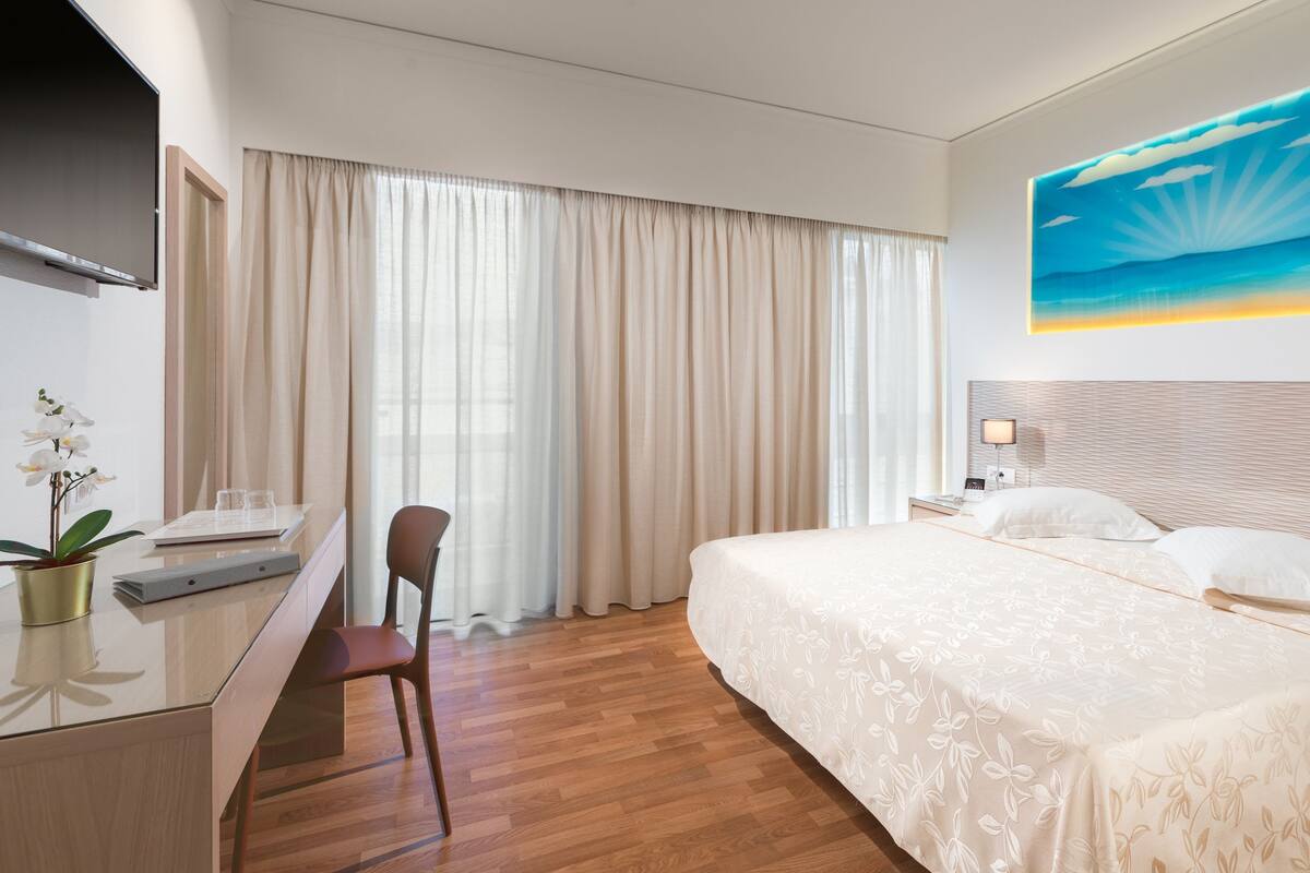A hotel room with a bed, a desk, a chair, a TV, and a painting of a sailboat on the wall.