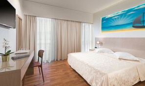 A hotel room with a bed, a desk, a chair, a TV, and a painting of a sailboat on the wall. - Alexandra Hotel (Kos)