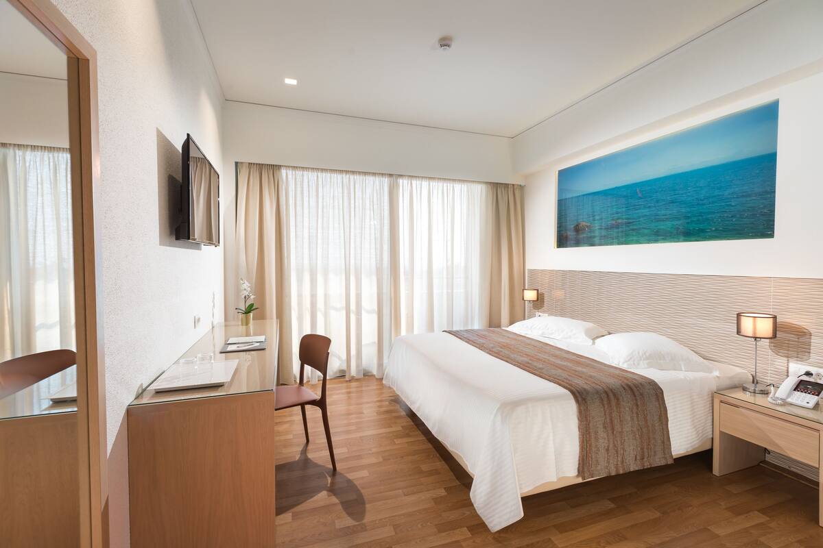 A hotel room with a large bed, a desk with a chair, a bedside table with a lamp, and a large window with curtains.