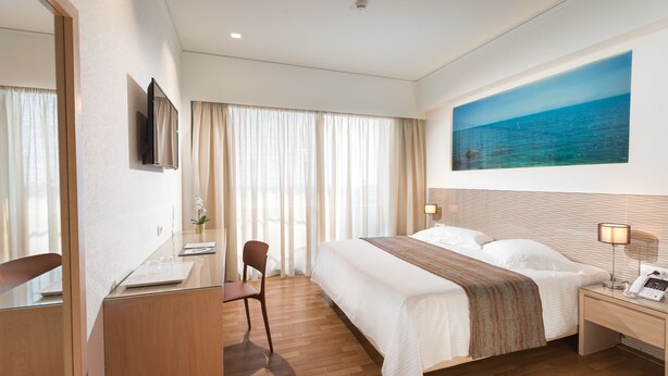 A hotel room with a large bed, a desk with a chair, a bedside table with a lamp, and a large window with curtains.