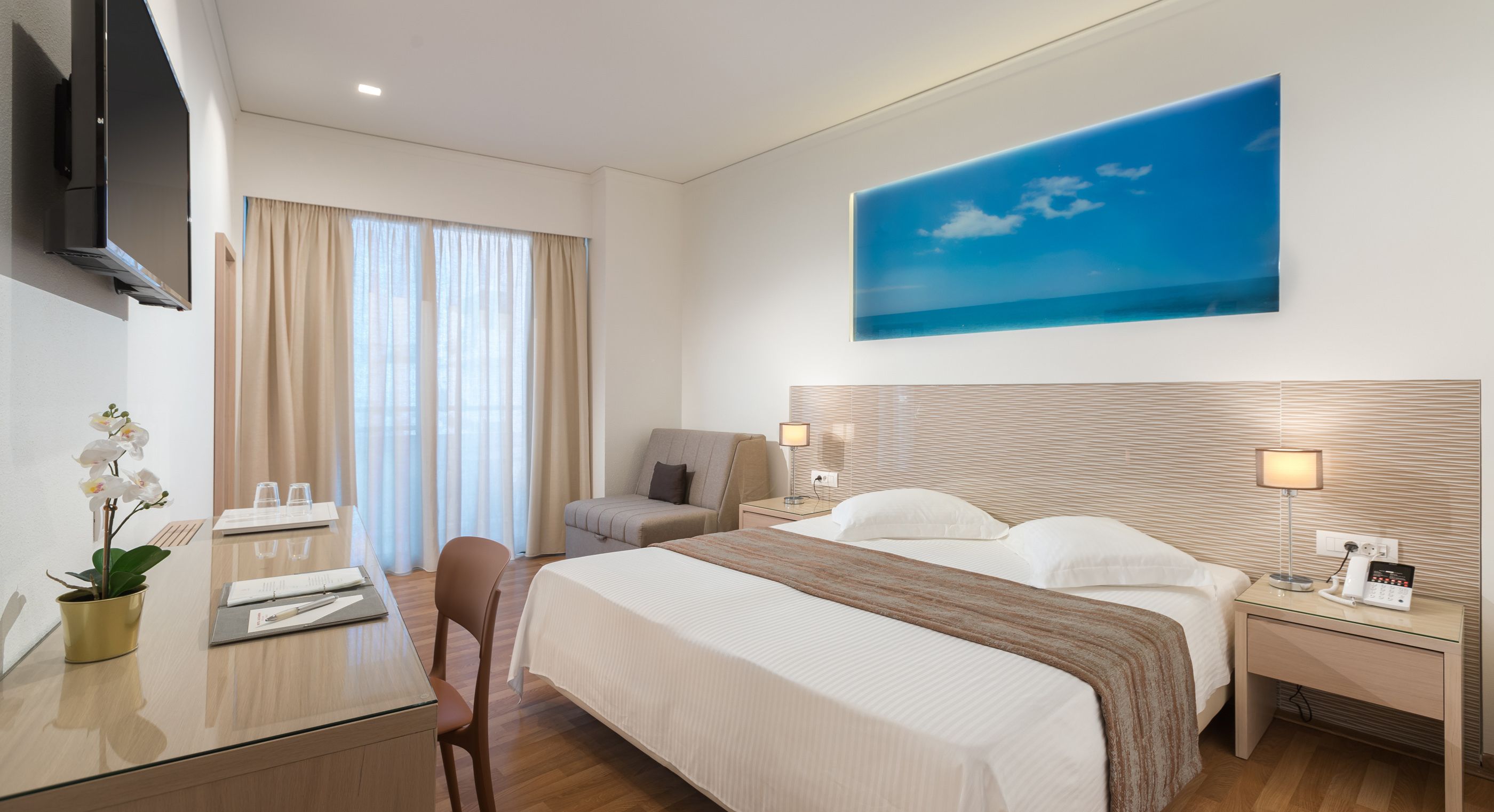 A hotel room with a bed, a desk, a chair, a TV, and a painting of a blue sky.