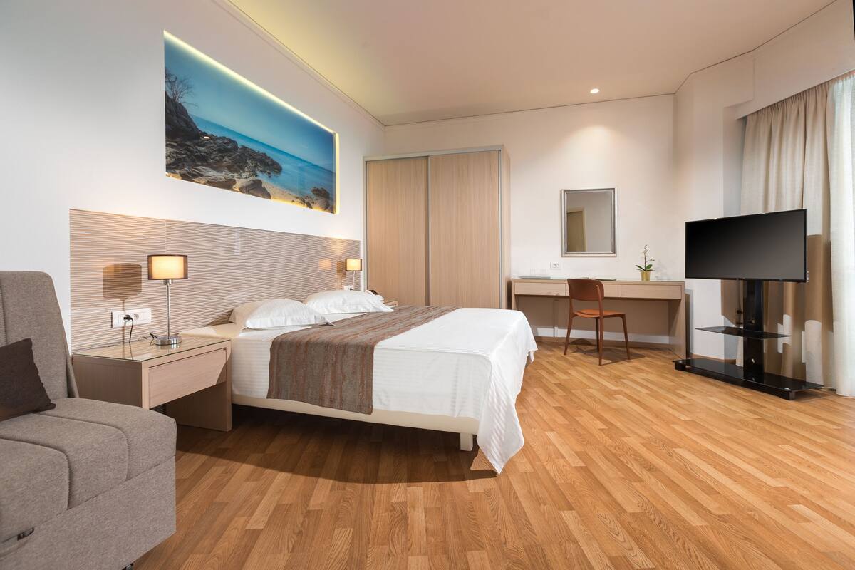A modern hotel room with a bed, a desk, a chair, a TV, and a painting of a beach.