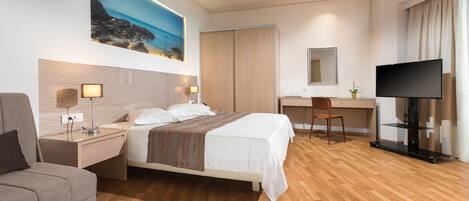 A modern hotel room with a bed, a desk, a chair, a TV, and a painting of a beach.