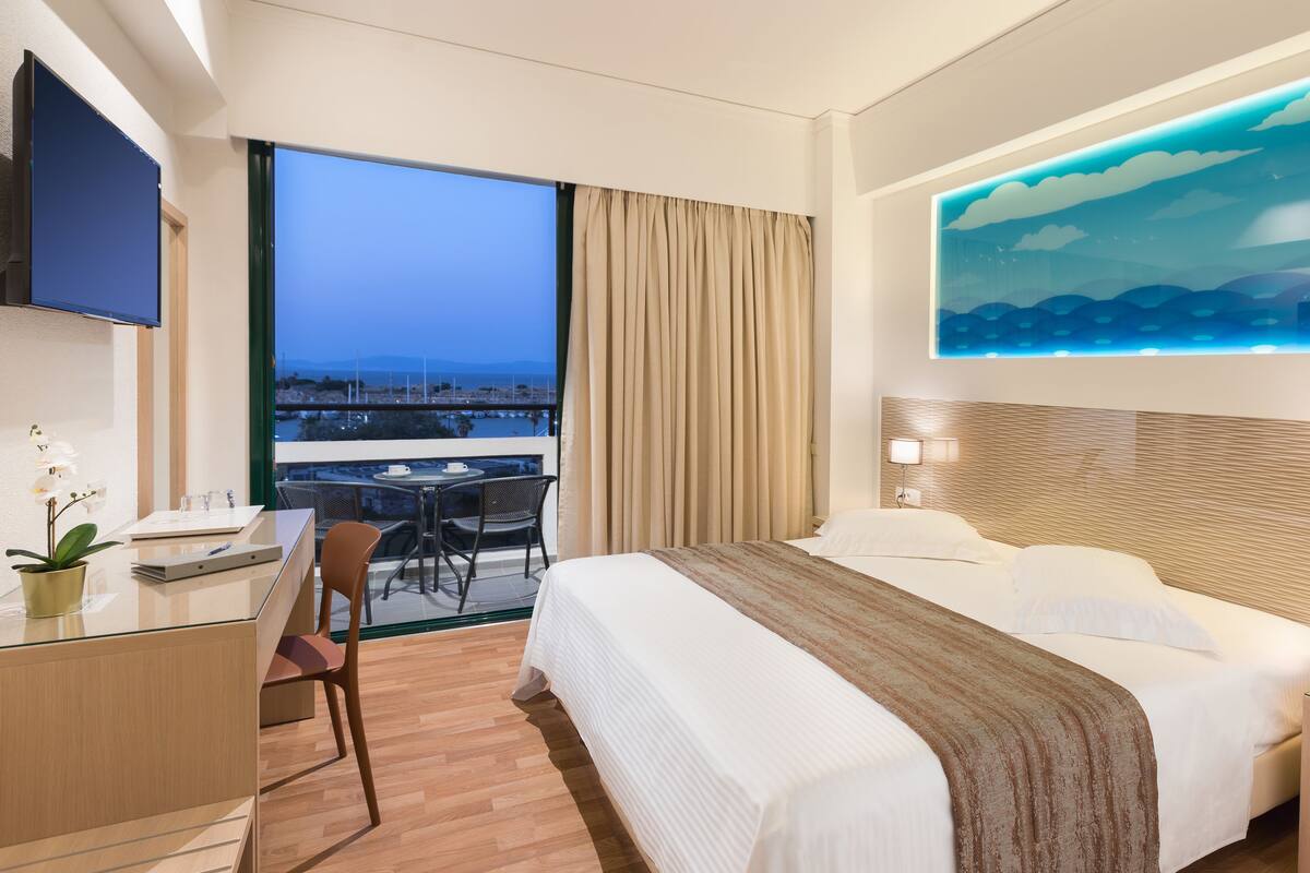 A hotel room with a large bed, a desk, a chair, a small table with a cup and saucer, a potted plant, a painting of a landscape, and a view of the sea through the window.