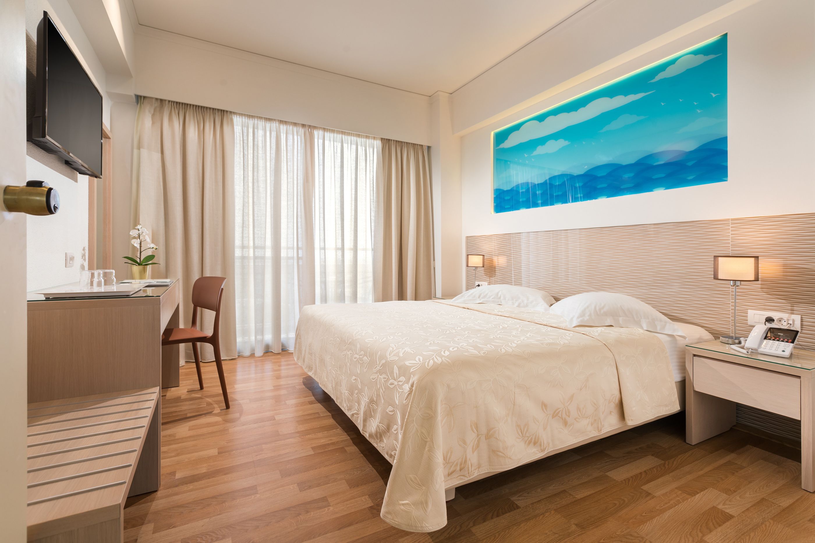 A hotel room with a large bed, a desk with a chair, a flat-screen TV, and a painting of a blue sky.