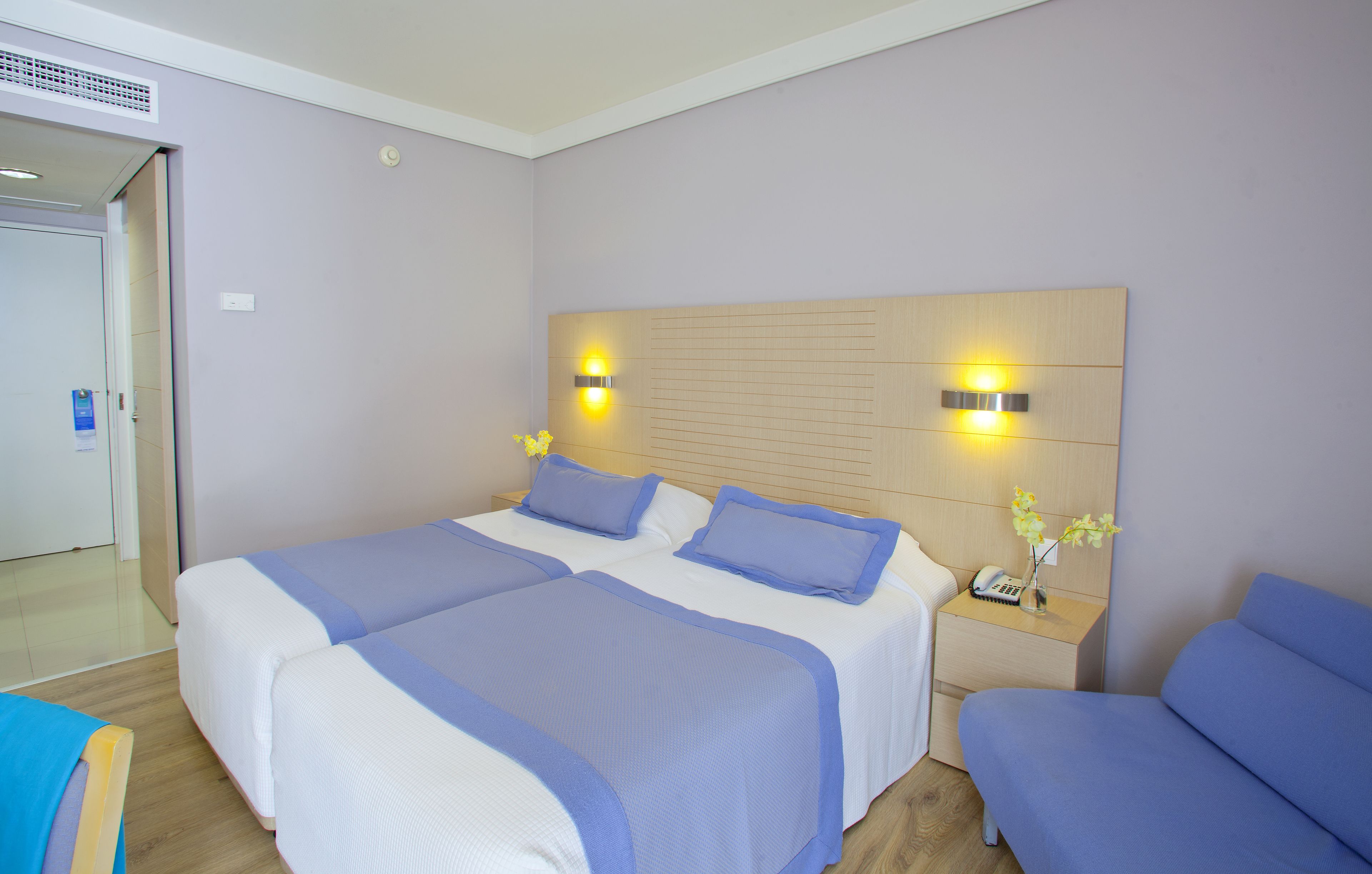 Habitación de hotel con dos camas, cabecera de madera con luces fijadas a la pared, mesita de noche con un teléfono y un florero, y una puerta a la izquierda.