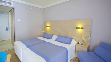 Habitación de hotel con dos camas, cabecera de madera con luces fijadas a la pared, mesita de noche con un teléfono y un florero, y una puerta a la izquierda.