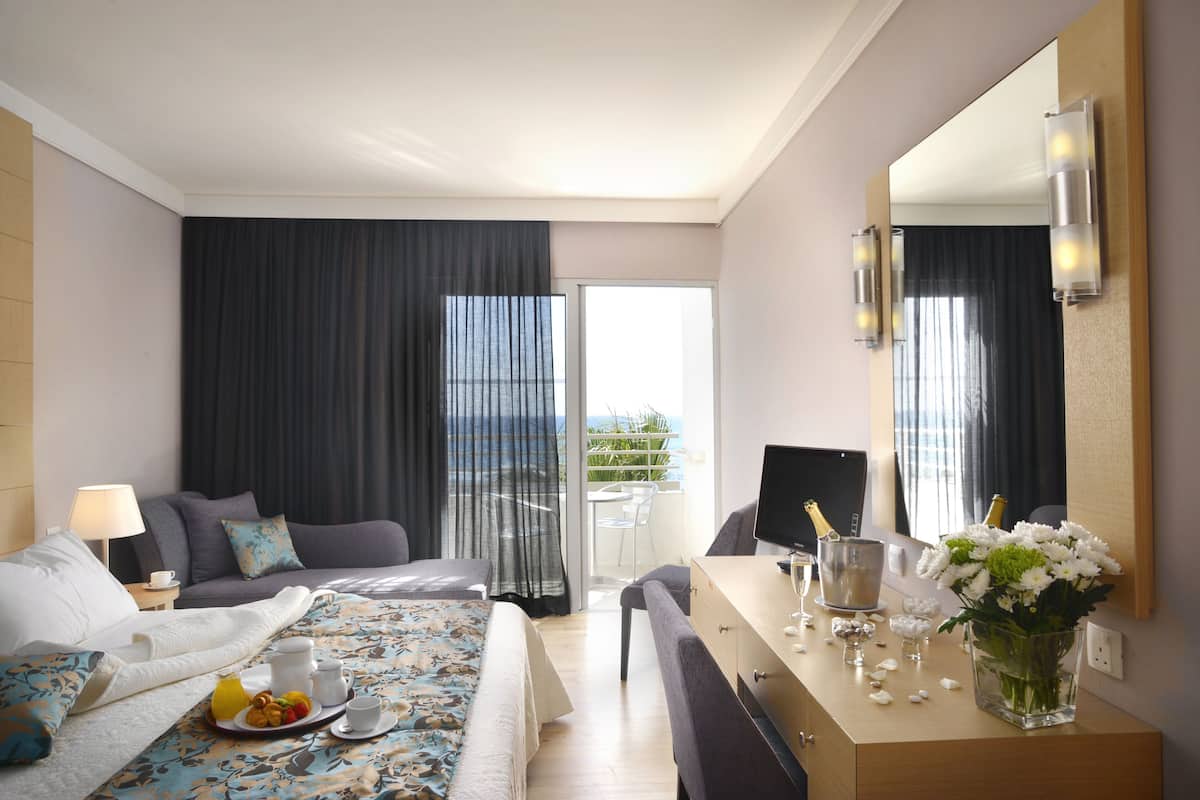 A hotel room with a bed, a desk with a computer, a chair, a small table with a tray of food, a vase of flowers, and a large mirror.