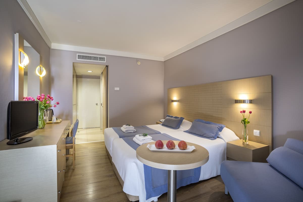 A hotel room with two beds, a desk with a computer, a small table with fruit, and a TV.