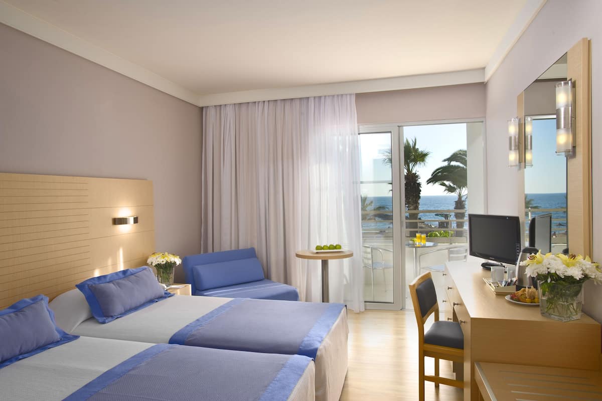 A hotel room with two beds, a desk with a computer, a chair, a small table with a vase of flowers, and a balcony with a view of the ocean.