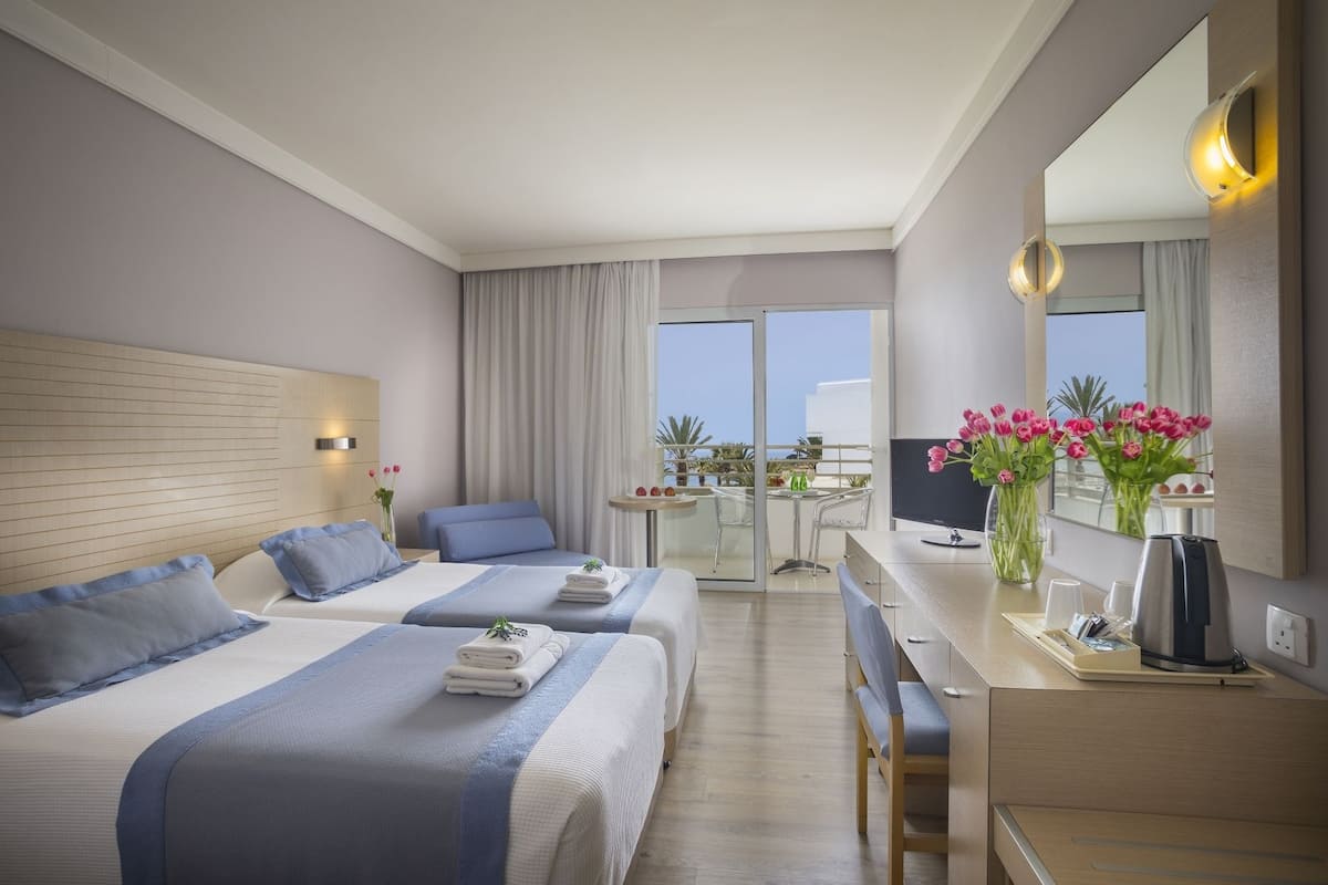 A hotel room with two beds, a desk, a chair, a small table, a mirror, and a balcony with a view.