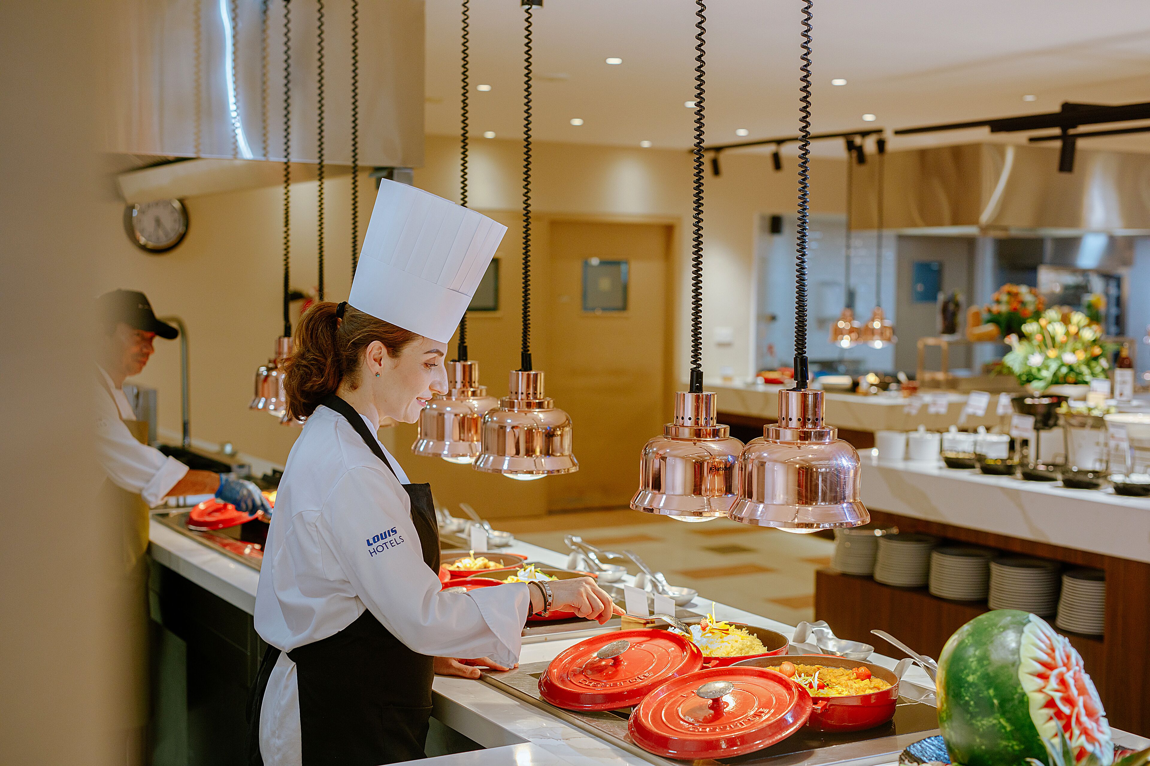 Una cocina de hotel con una disposición tipo bufé, que incluye diferentes estaciones de comida y una sandía decorativa de gran tamaño.