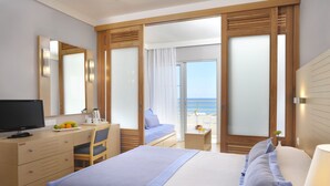 Habitación de hotel con cama, escritorio con computadora, silla, pequeña mesa con lámpara y vista al mar a través de una puerta de cristal.