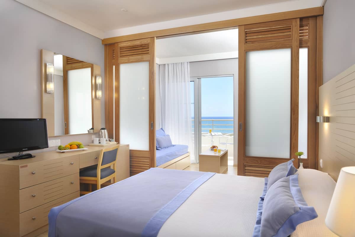 A hotel room with a bed, a desk with a computer, a chair, a small table with a lamp, and a view of the sea through a glass door.