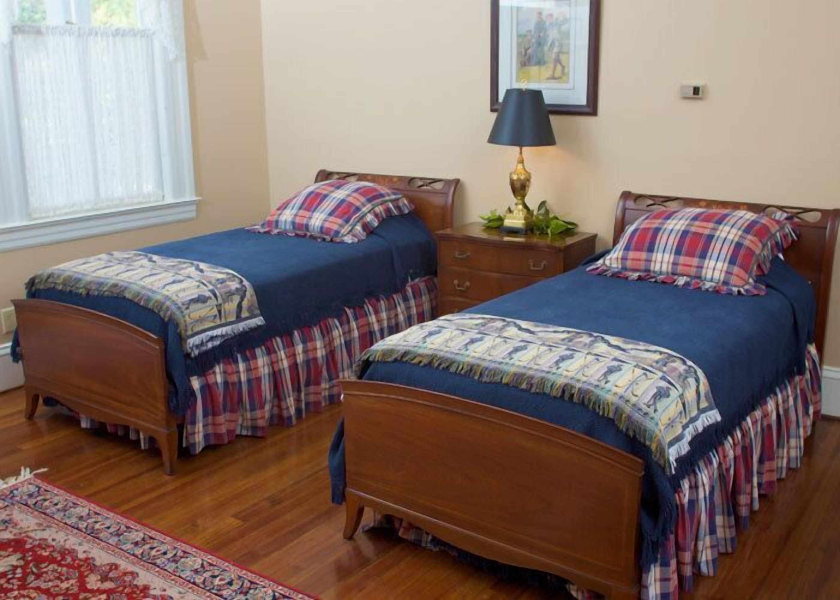 A bedroom with two wooden beds, a nightstand with a lamp, a framed picture on the wall, and a window with sheer curtains.