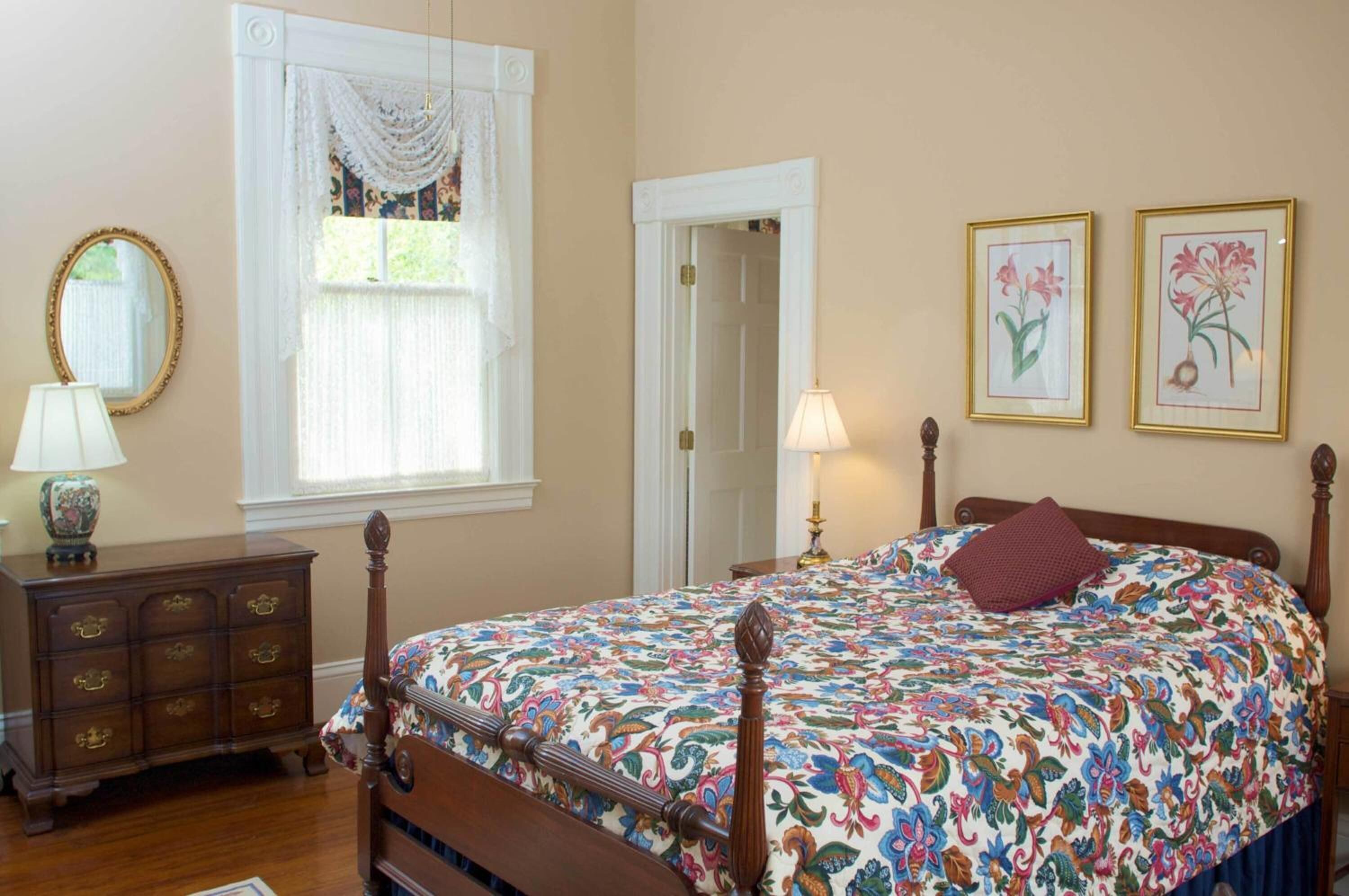 A bedroom with a floral bedspread, a wooden dresser, a mirror, and two framed floral paintings on the wall.