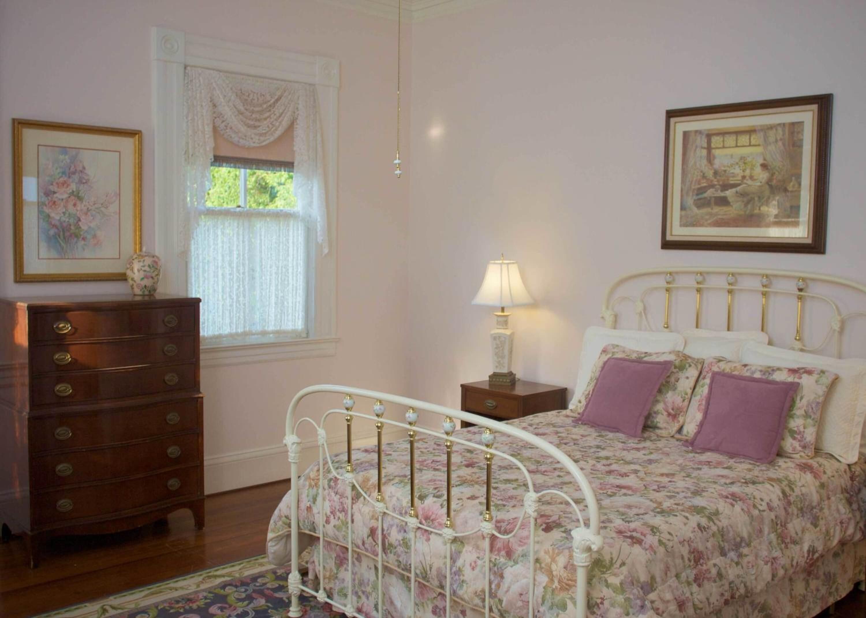 A bedroom with a bed, dresser, window, and paintings.