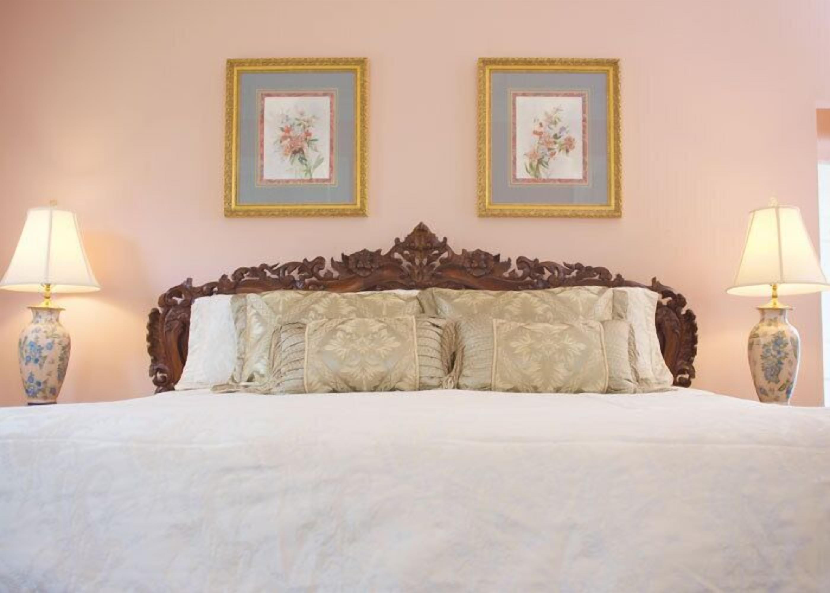Uma cama com cabeceira de madeira, duas pinturas florais emolduradas na parede e duas luminárias de cada lado.