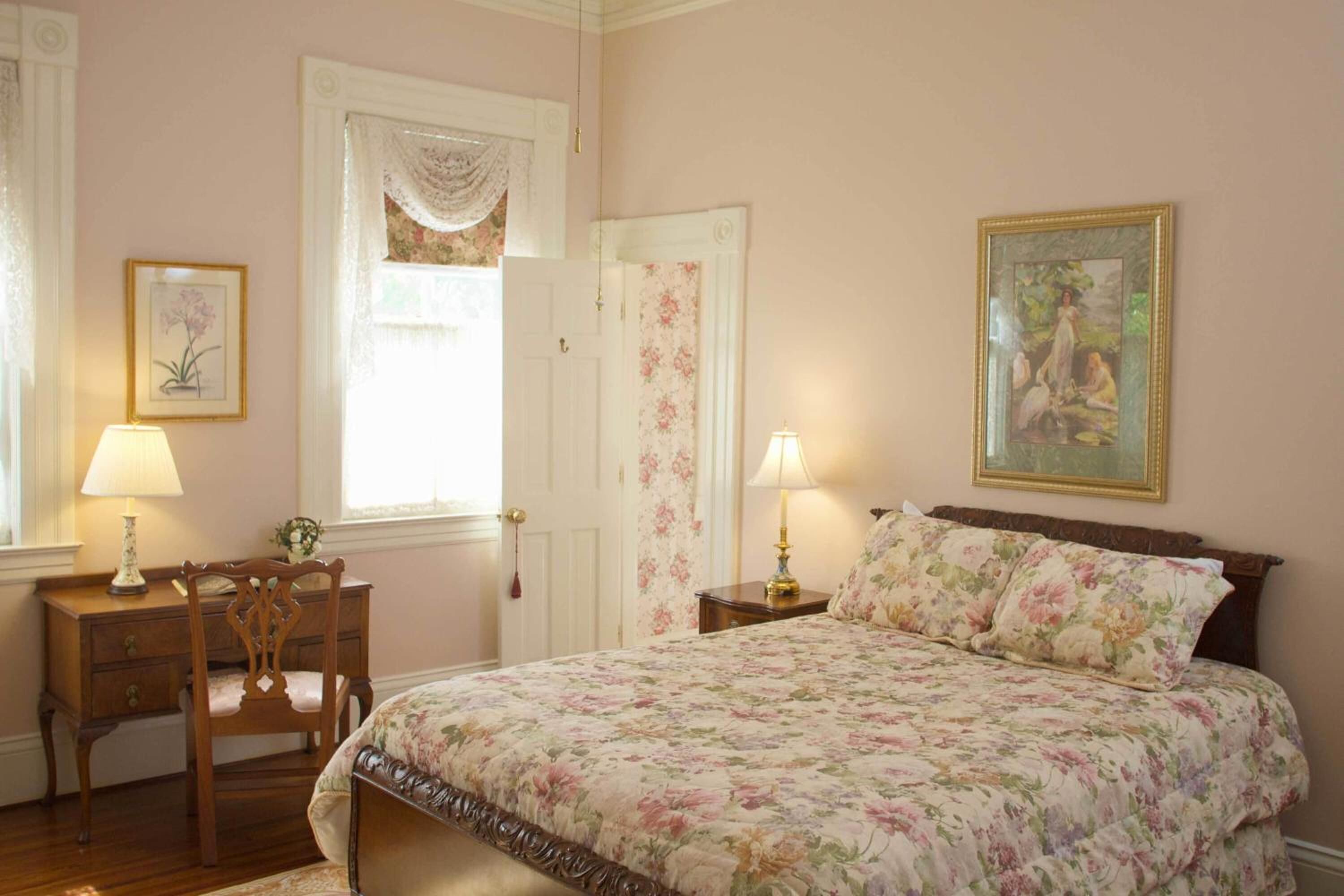A bedroom with a floral bedspread, a wooden chair, a desk with a lamp, and a painting on the wall.