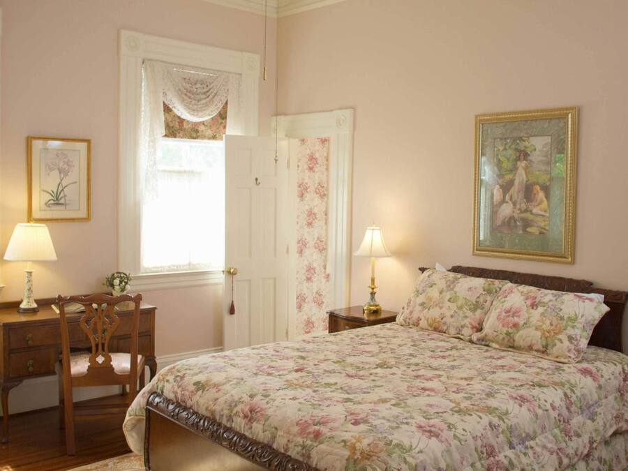 A bedroom with a floral bedspread, a wooden chair, a desk with a lamp, and a painting on the wall.