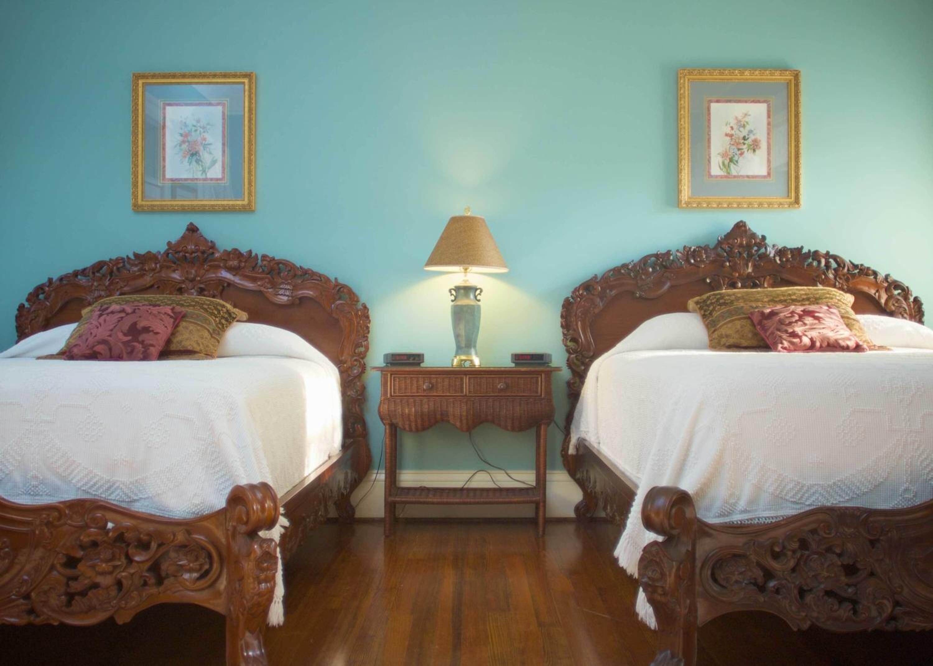 A room with two ornate wooden beds, a small nightstand with a lamp, and floral wall art.