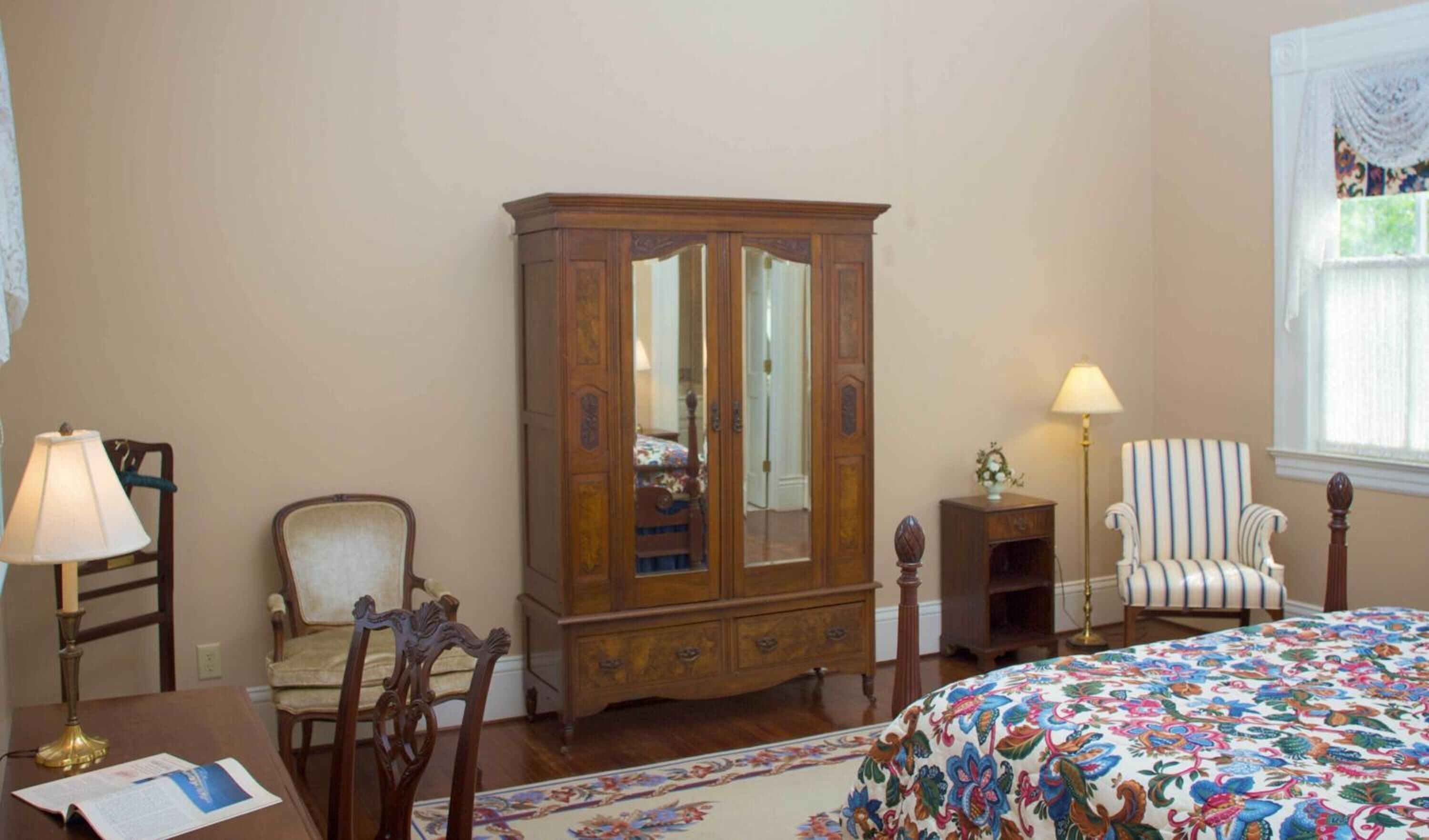 A room with a large wooden armoire, a striped armchair, a bed with a floral bedspread, a desk with a lamp, and a window with a curtain.