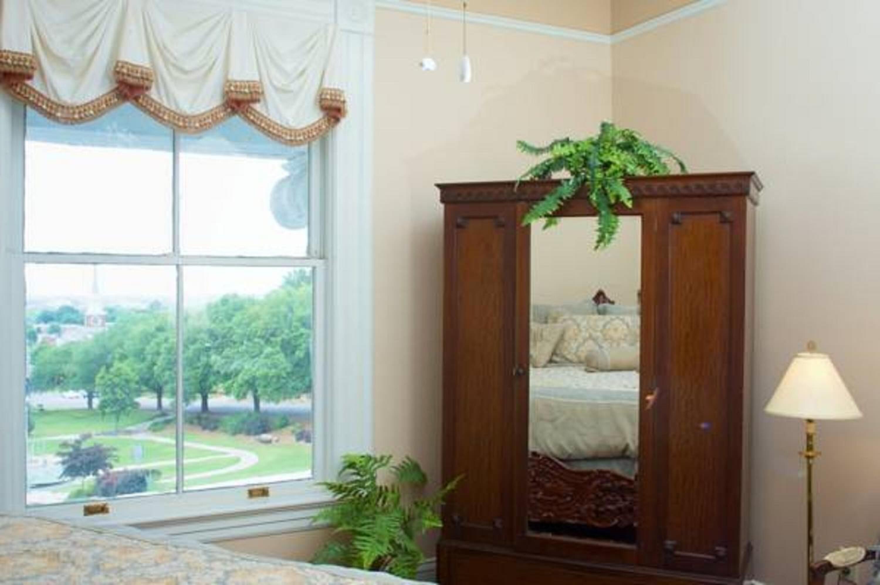 A bedroom with a large window, a wooden wardrobe, a bedside lamp, and a view of a park.