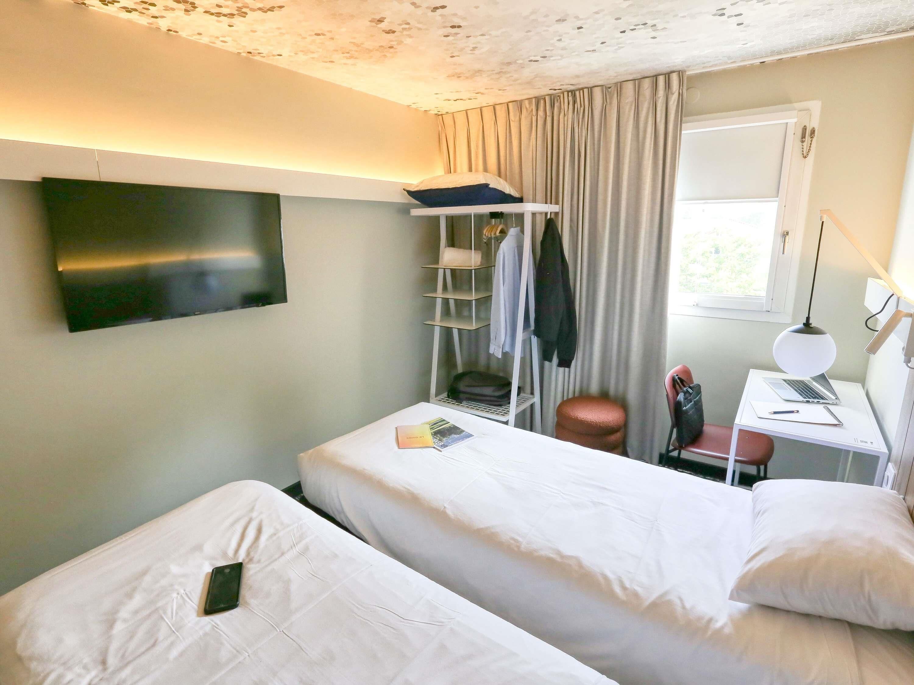 A hotel room with two beds, a TV, a desk with a laptop, a chair, a shelf with clothes, and a window.