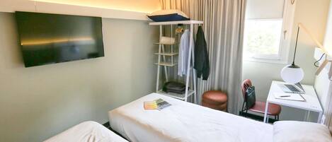 A hotel room with two beds, a TV, a desk with a laptop, a chair, a shelf with clothes, and a window.