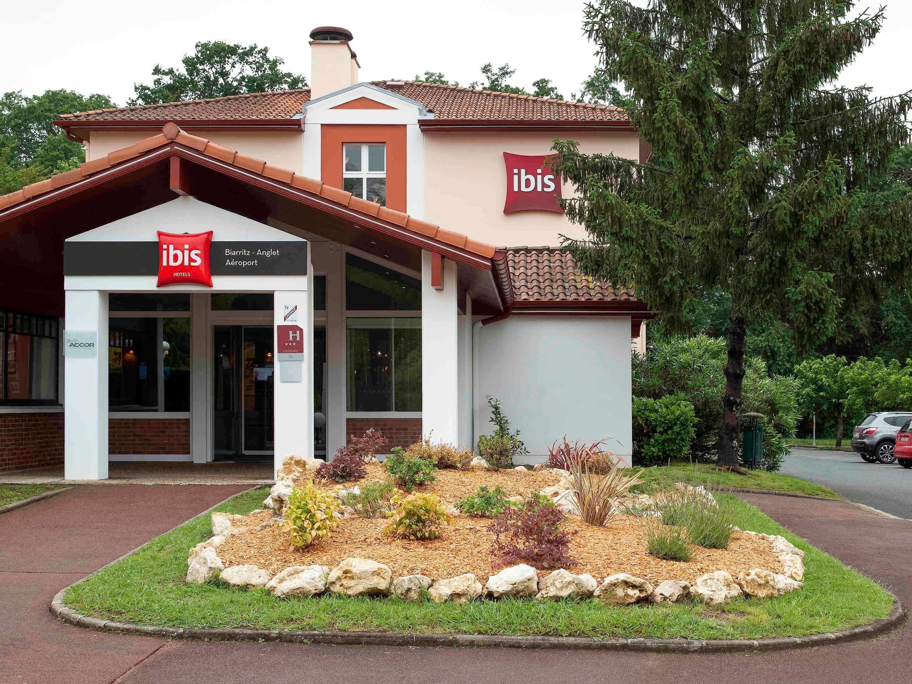 Un hotel con fachada roja y blanca, un cartel que indica que es un hotel ibis, y un jardín ajardinado frente a él.