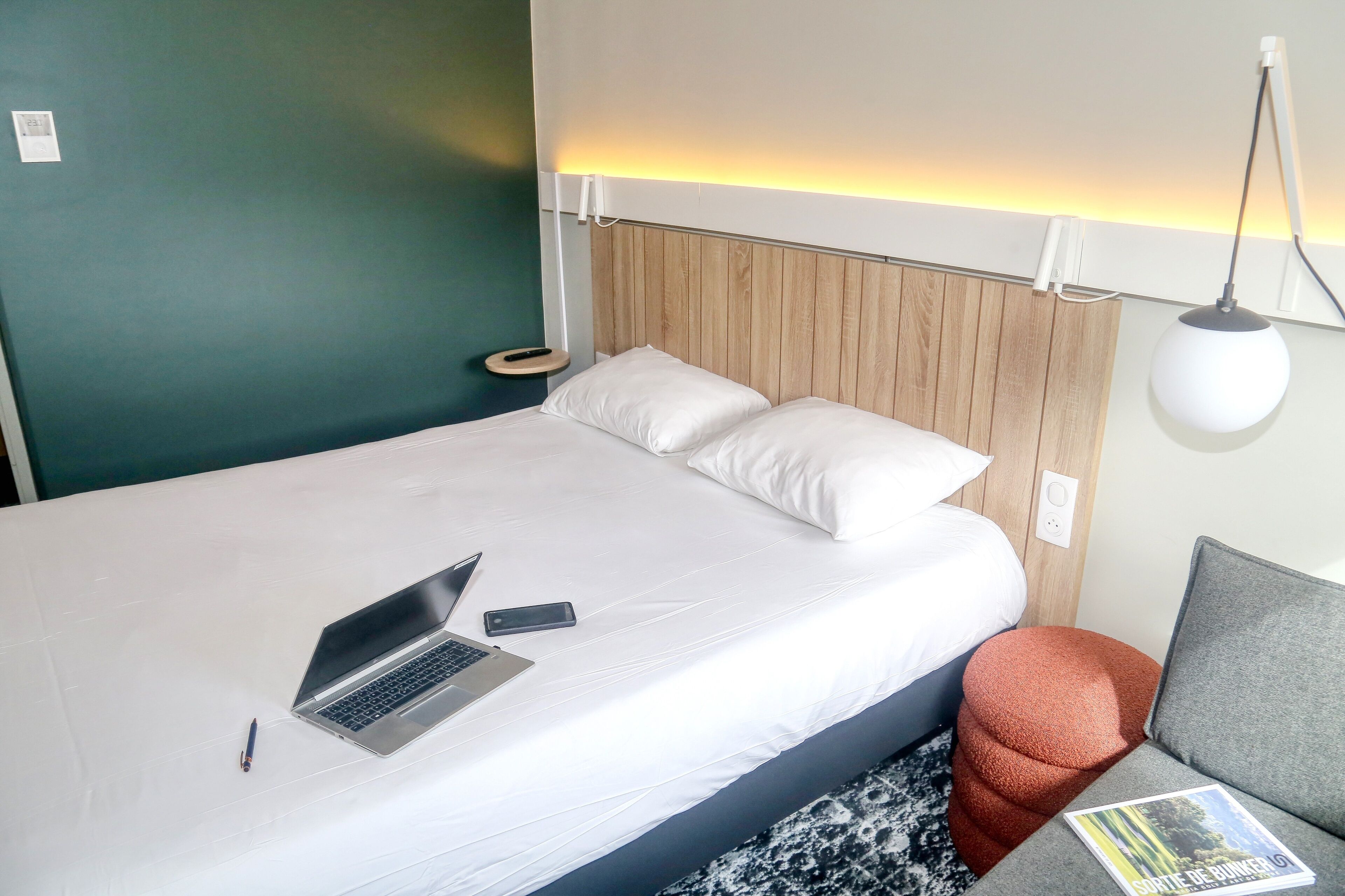 En pænt redt seng med en bærbar computer og en fjernbetjening, et sengebord med et magasin, og en lampe monteret på væggen.
