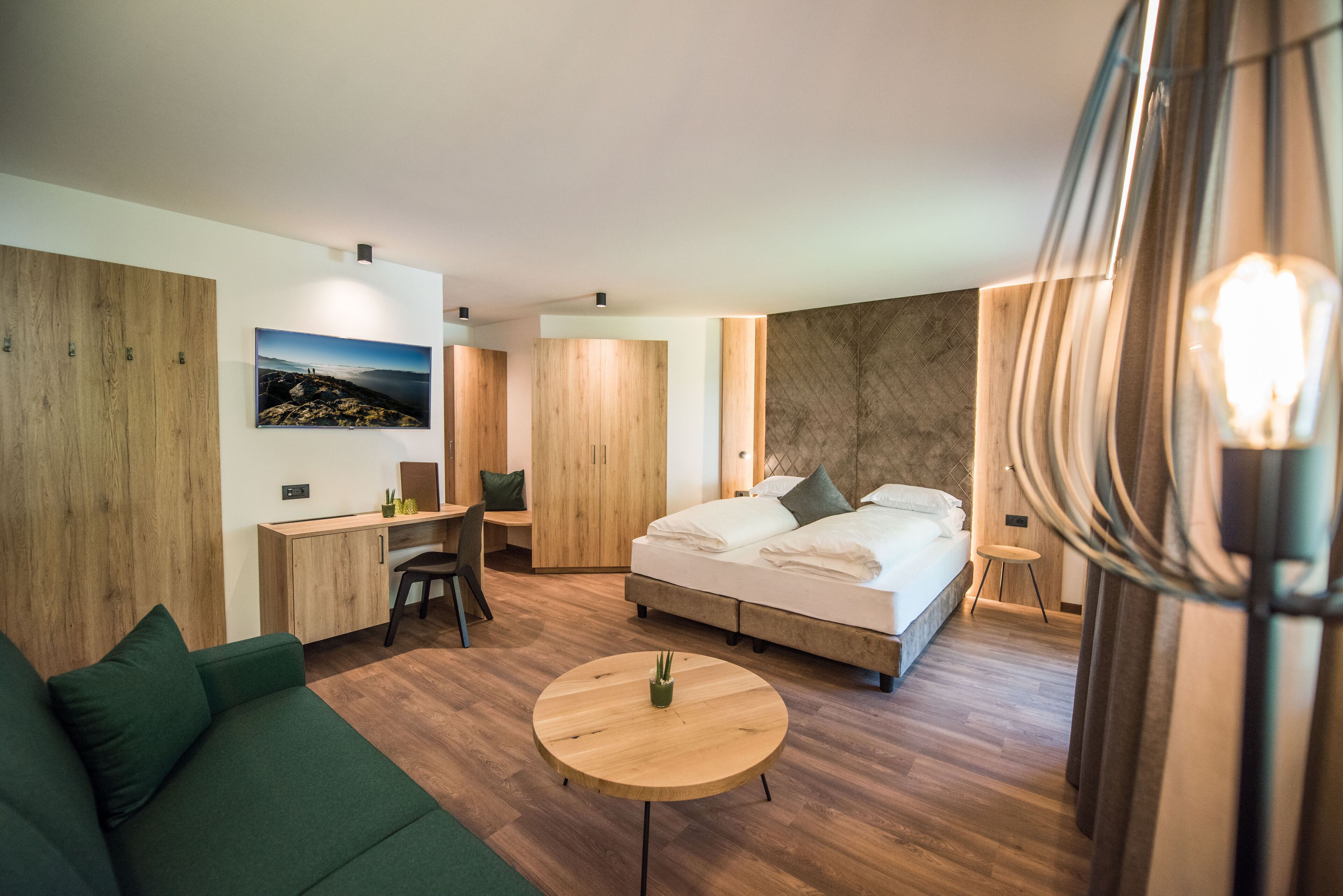 Quarto de hotel moderno com uma cama, uma escrivaninha, um sofá verde e uma mesa redonda de madeira.