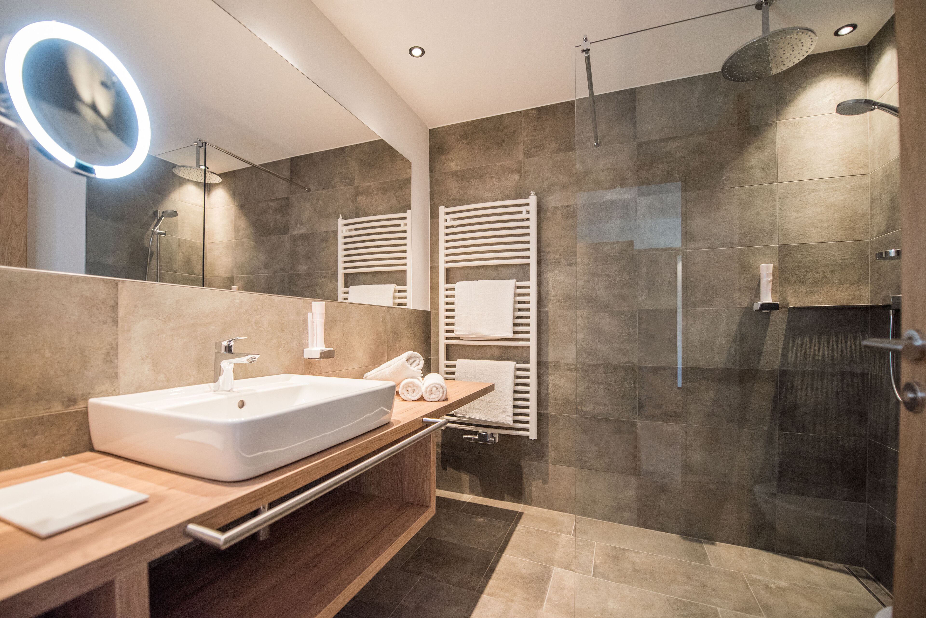 A modern bathroom with a large sink, a mirror with a light, a shower, and towel racks.