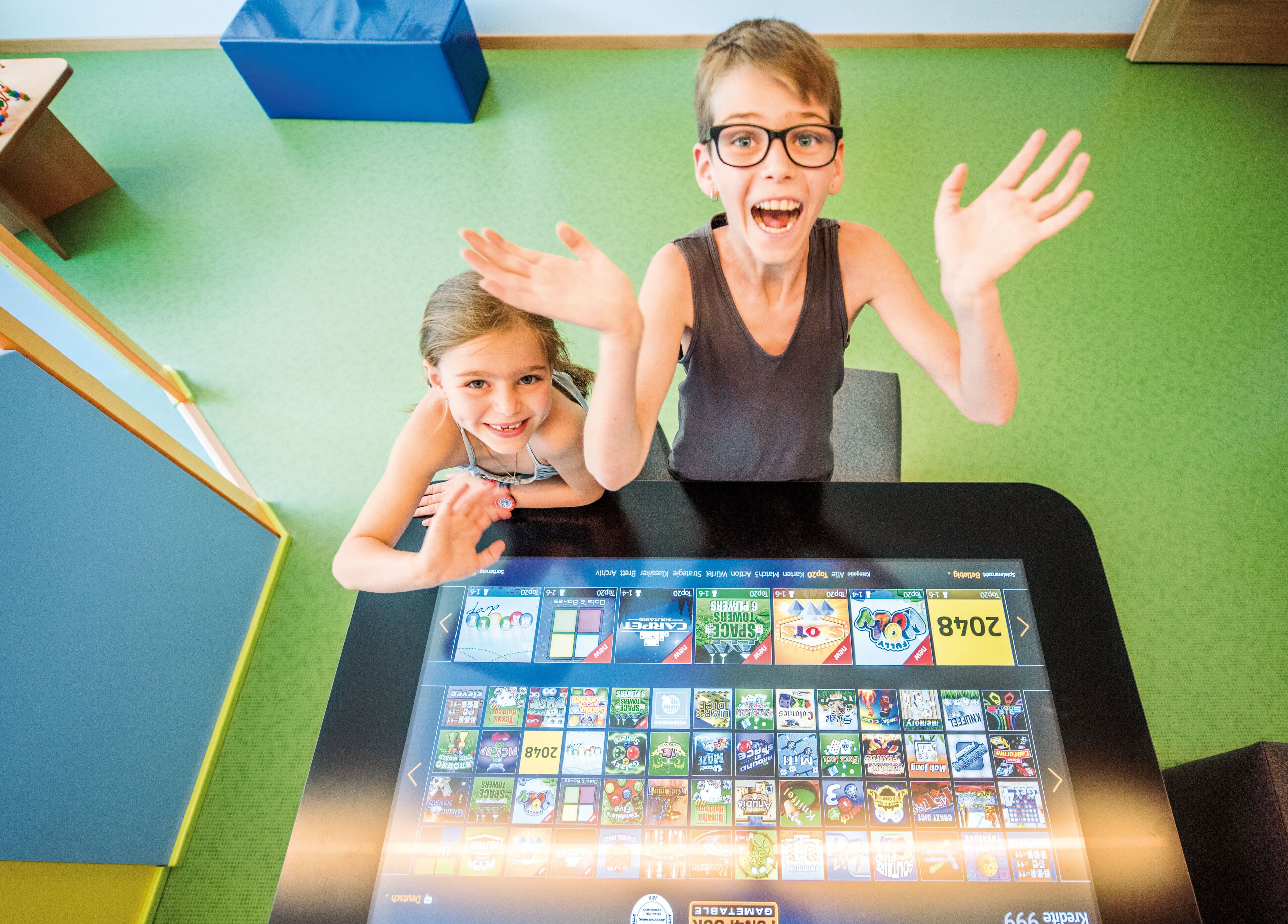 A room with a green carpet, a black table displaying a digital screen with game icons, and a blue storage box in the background.