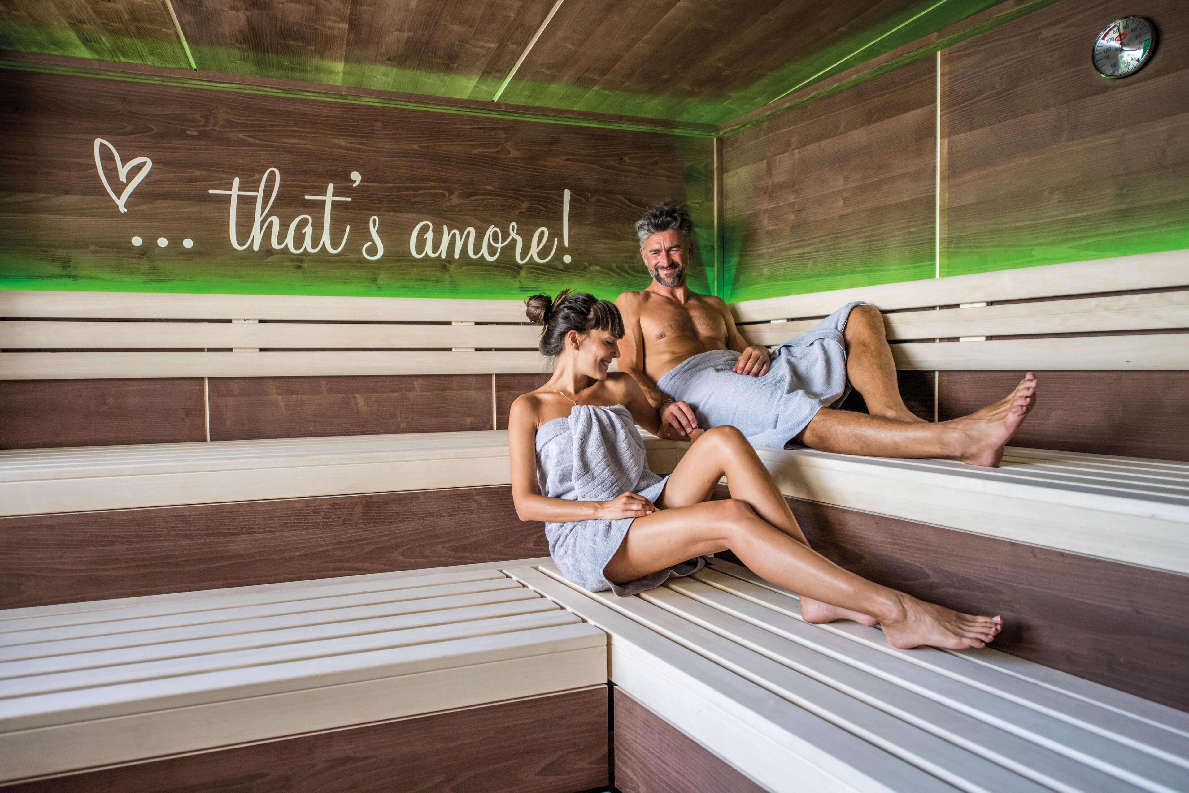 Una pareja sentada en bancos de madera en una habitación con una pared verde y un letrero que dice 'that's amore!'.