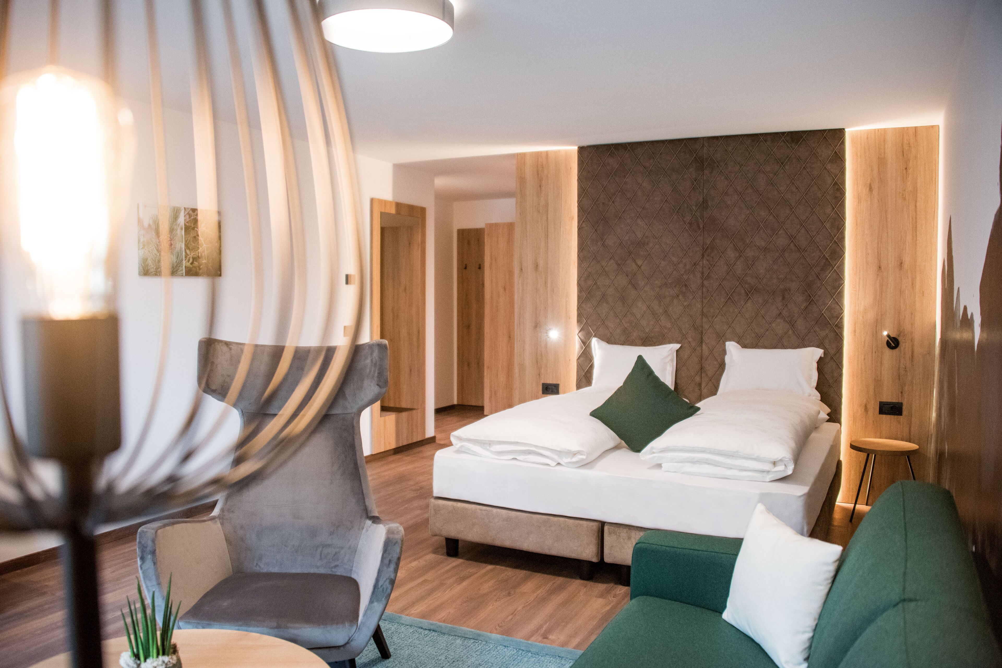 Une chambre d’hôtel moderne avec un grand lit, un canapé vert, un fauteuil gris et un mur décoratif en bois.