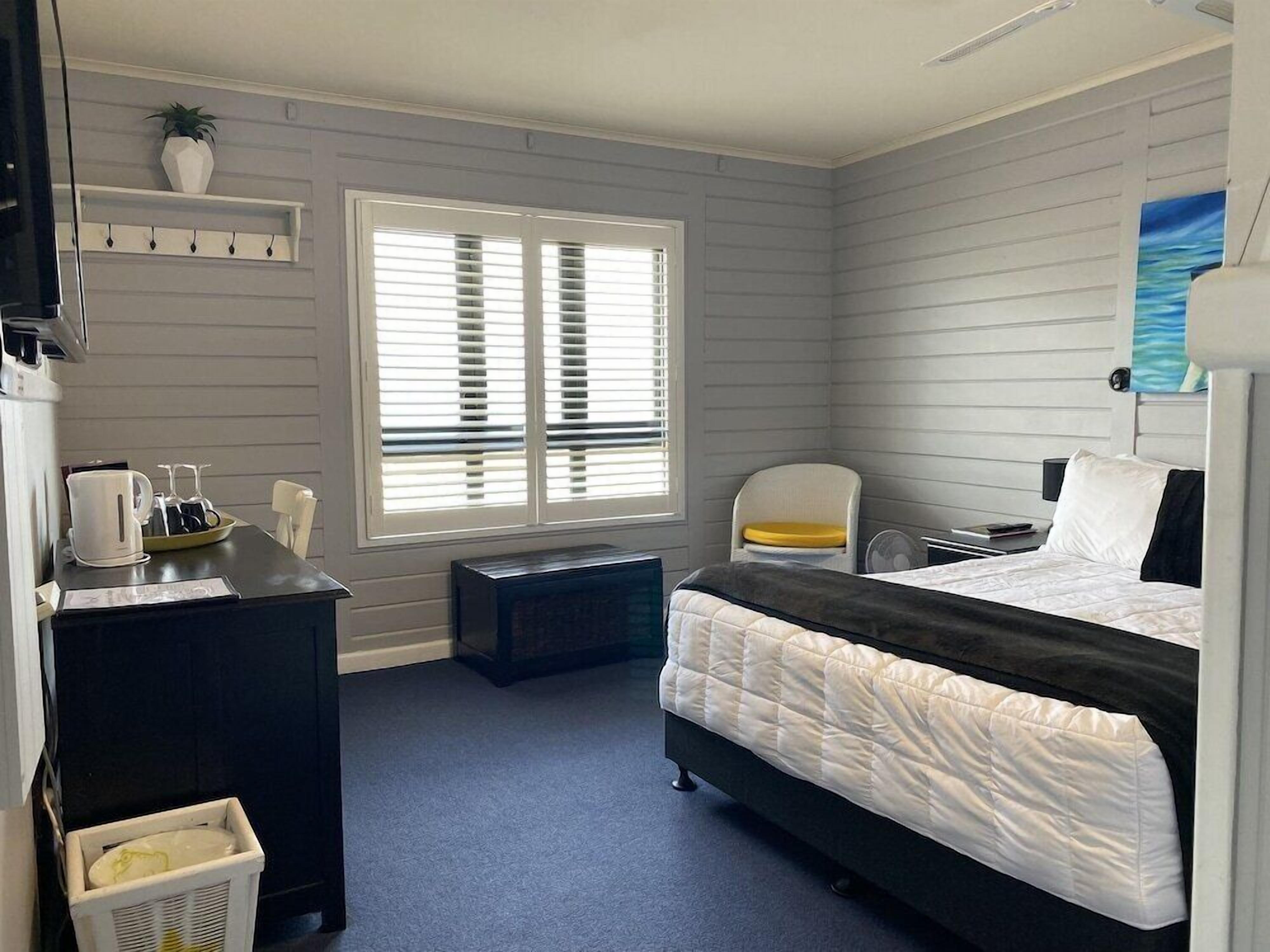 A hotel room with a bed, a desk, a chair, a small table, a window with blinds, and a wall-mounted shelf with a plant.