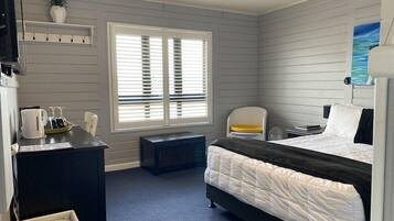 A hotel room with a bed, a desk, a chair, a small table, a window with blinds, and a wall-mounted shelf with a plant.