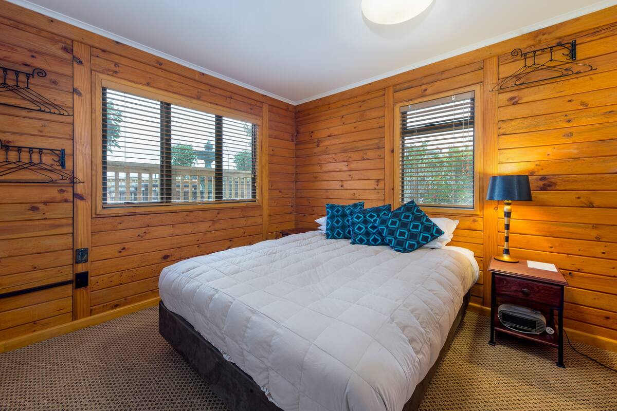 Une chambre avec des murs en bois, un lit avec une literie blanche, deux fenêtres équipées de stores, une table de chevet avec une lampe et un petit radiateur.