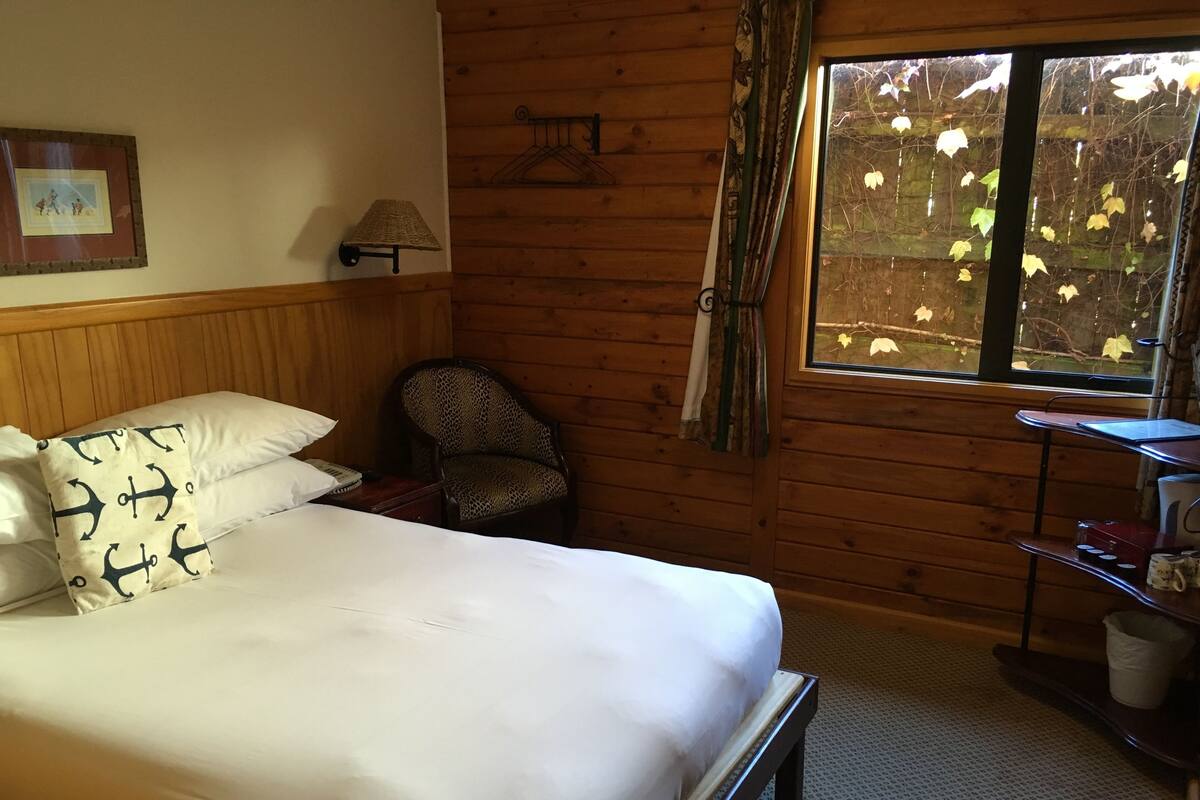 Une pièce de cabane en bois comprenant un lit, une chaise, une petite table et une fenêtre donnant sur la forêt.