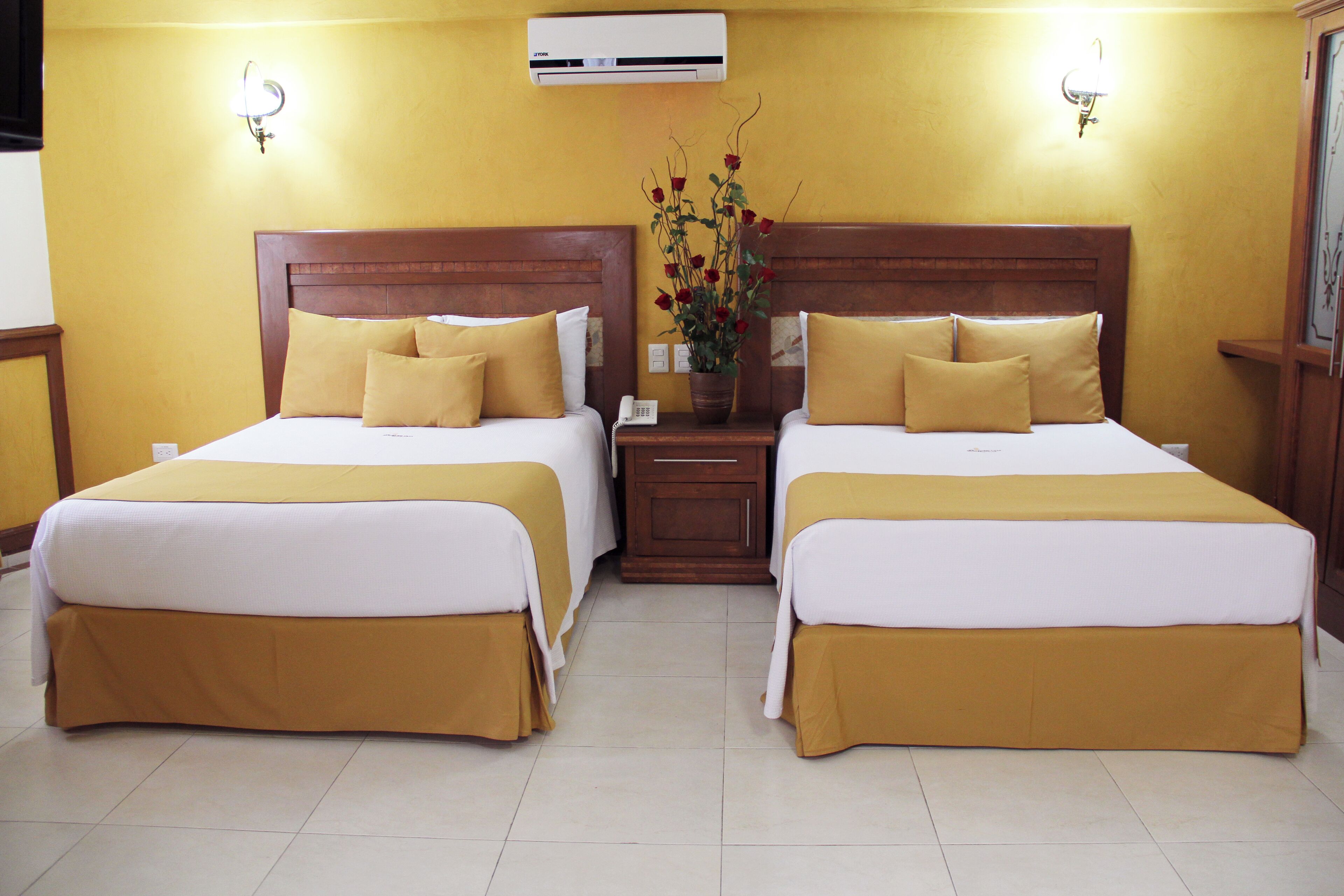 Habitación de hotel con dos camas, cada una con cabecero de madera, ropa de cama blanca y almohadas a juego. Se encuentra una mesita de noche con un teléfono y un florero con flores entre las camas.