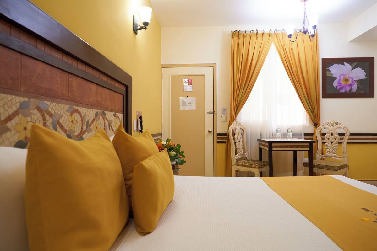 A hotel room with a bed, two chairs, a small table, a flower painting, and a door with a sign.