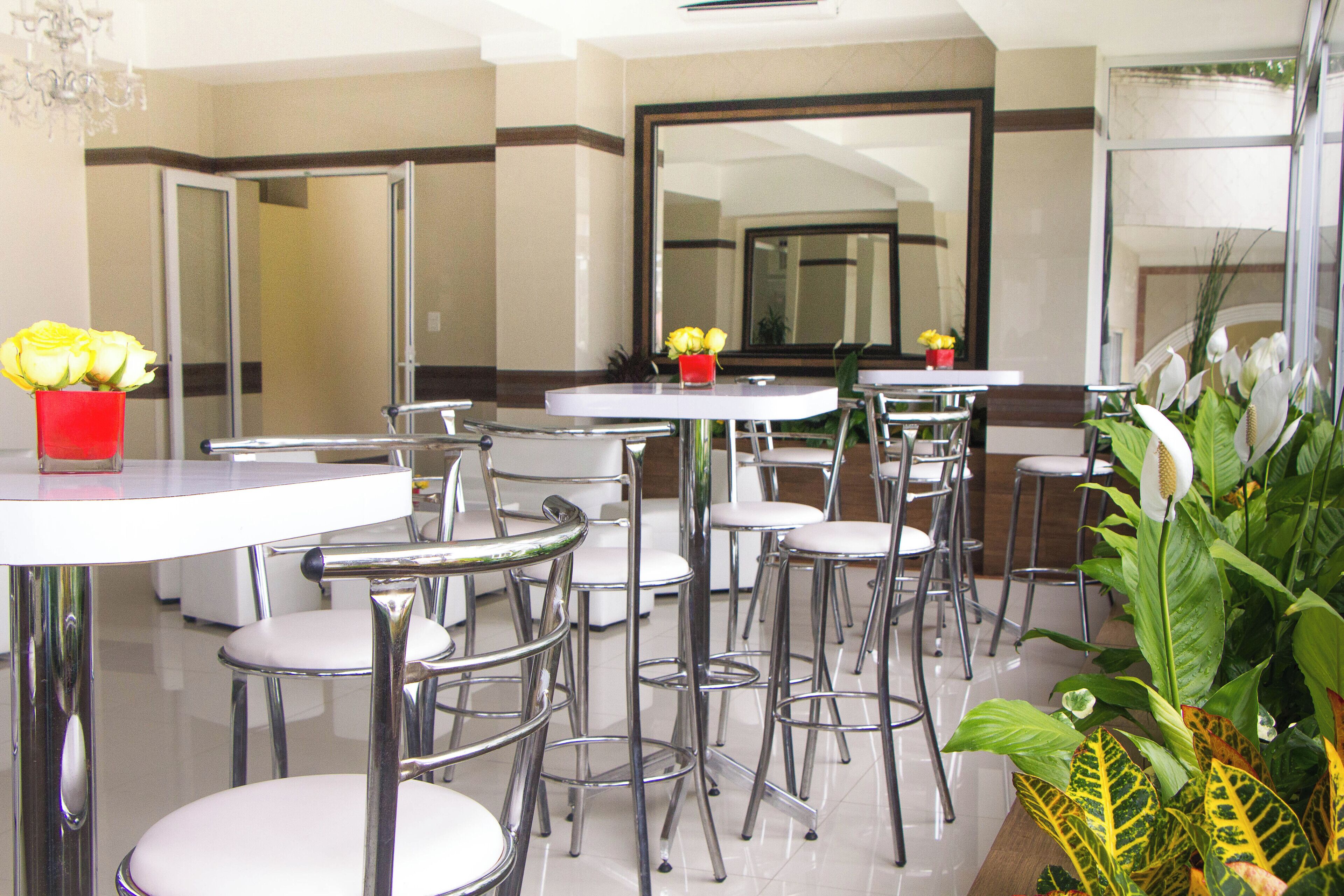 A modern hotel dining area with round tables and metal chairs, a red vase with yellow flowers, and a large mirror on the wall.