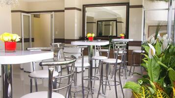 A modern hotel dining area with round tables and metal chairs, a red vase with yellow flowers, and a large mirror on the wall.