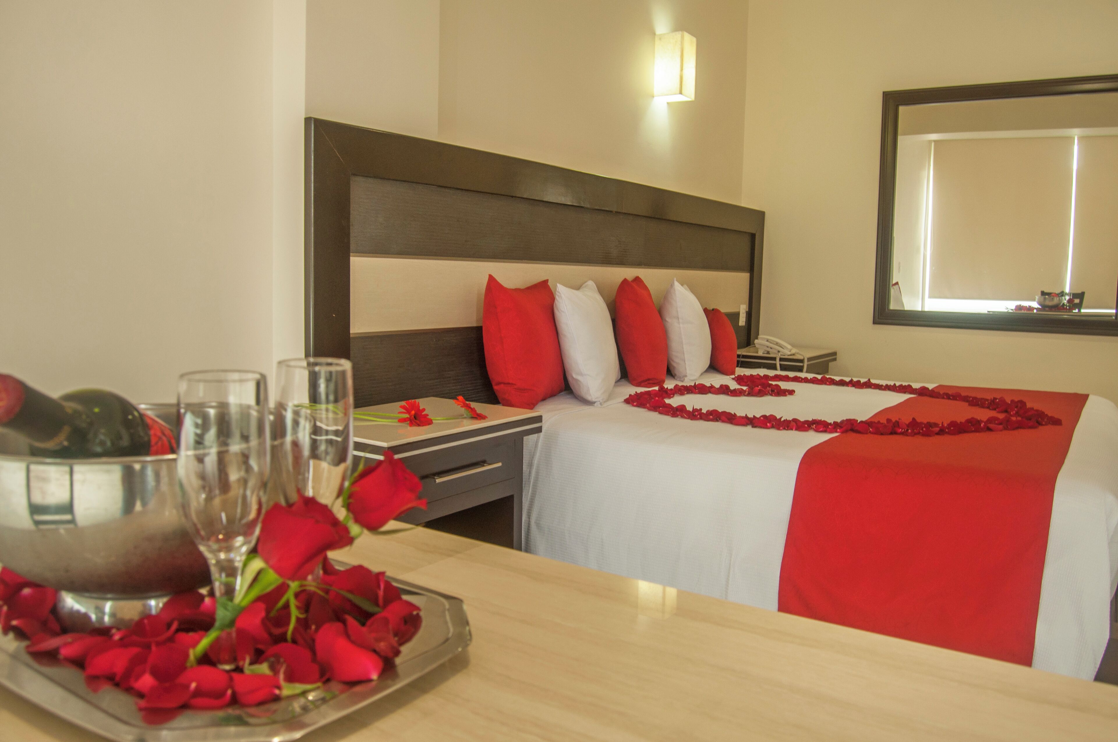 Una habitación de hotel con cama, una mesita de noche con un florero de rosas rojas y un minibar.