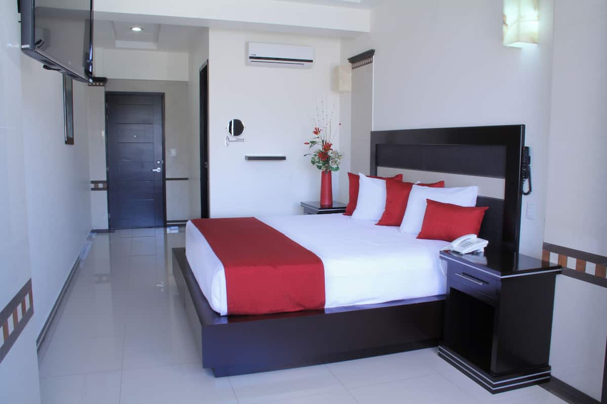 A hotel room with a large bed, a bedside table with a phone, a red vase with flowers, and a door leading to another room.