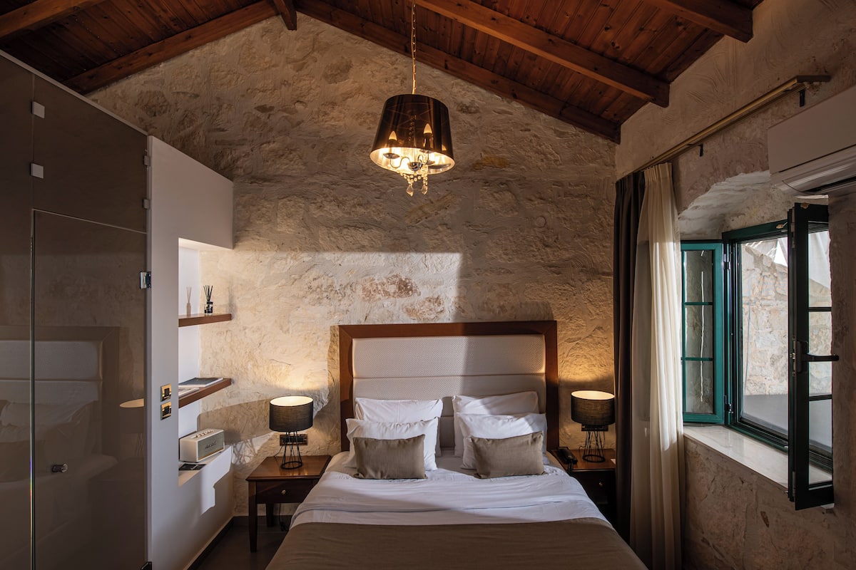 A bedroom with a stone wall, a wooden ceiling, a bed with white linens, two bedside tables with lamps, and a window with curtains.