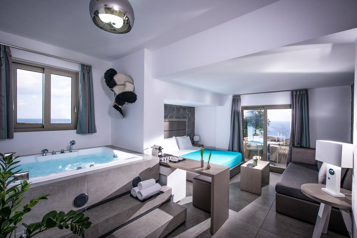 A modern hotel room with a large bathtub, a bed with a view, a small desk, and a seating area.