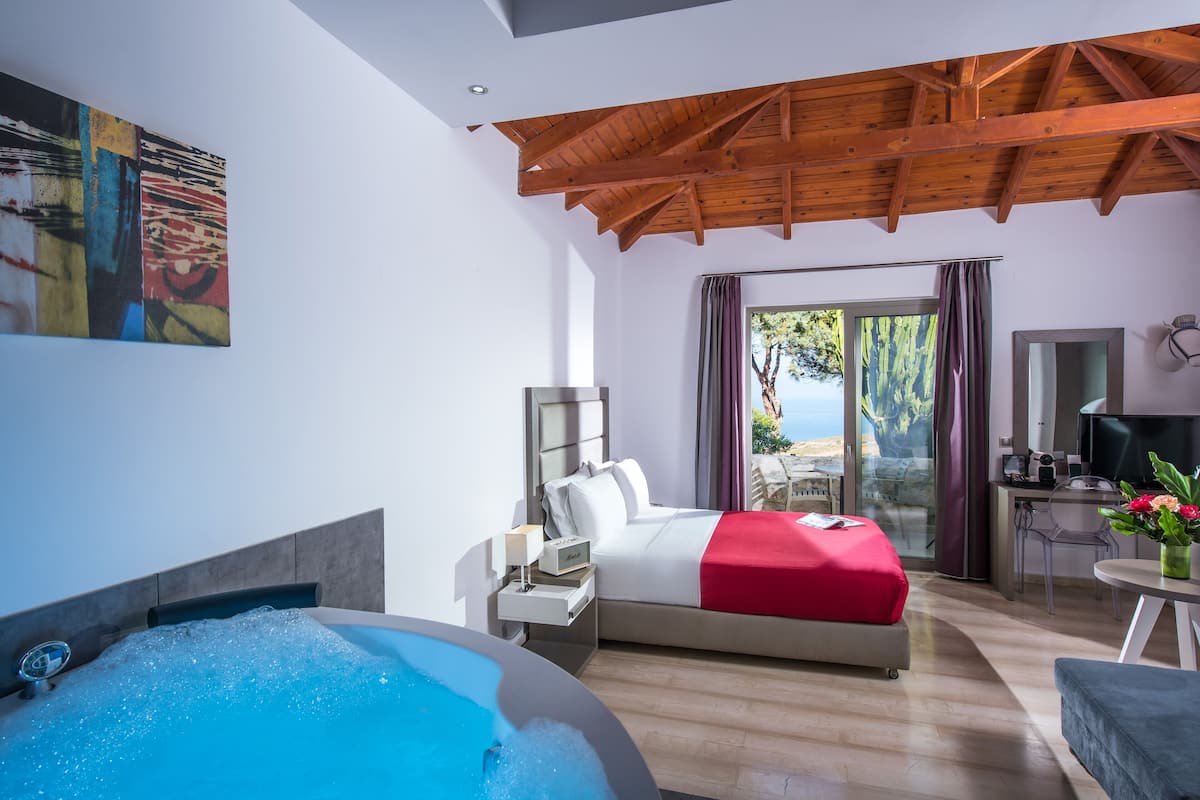 A modern hotel room with a large bed, a sitting area, a desk, and a view of the outdoors.